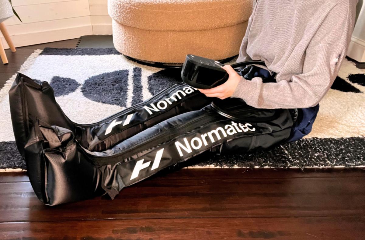 woman sitting on black and white rug in black normatec boots, holding the remote that controls the device