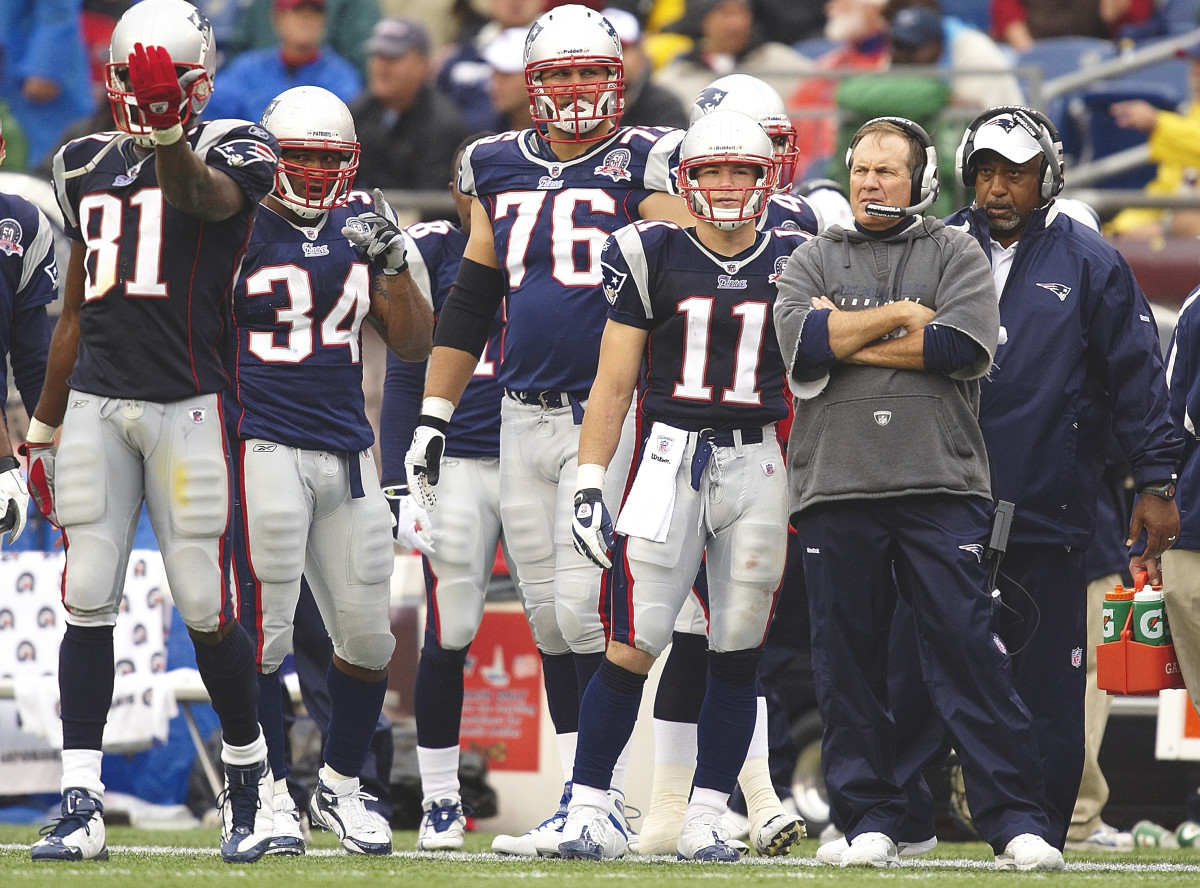 Bill Belichick stands on the sideline with his arms crossed next to a group of players