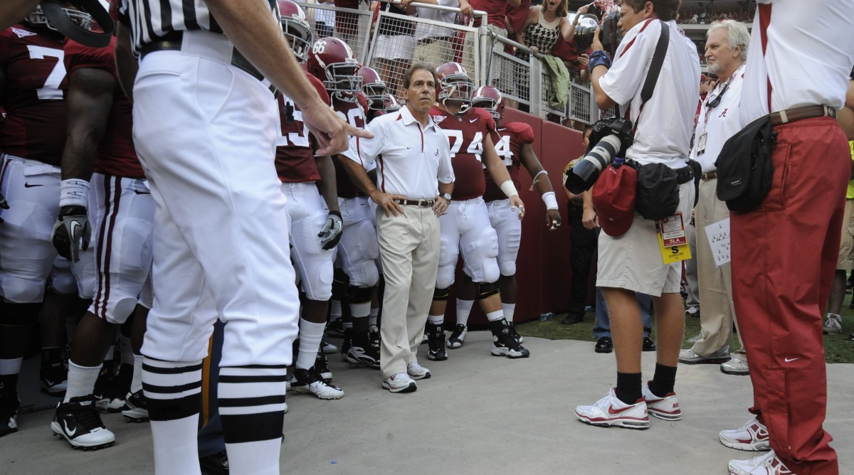 Alabama head coach Nick Saban looks on while standing with his hands on his hips in front of a group of his players.