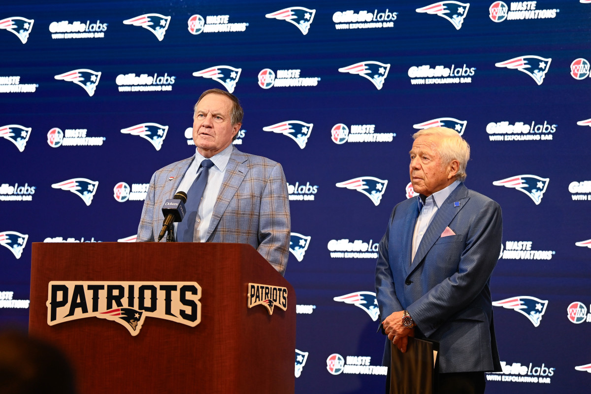 Bill Belichick stands talking at a podium with a microphone as Robert Kraft stands next to him