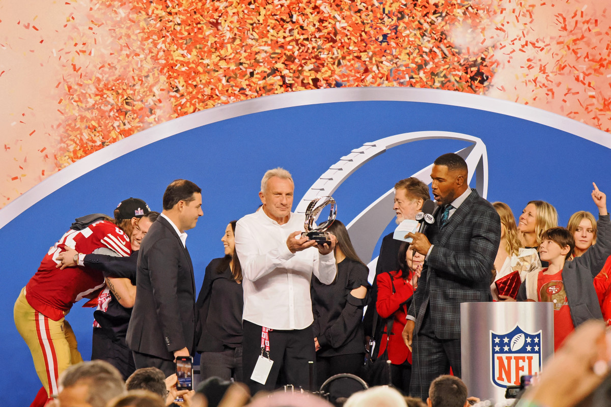 Joe Montana holds a trophy up while 49ers players and FOX commentators stand on stage with him